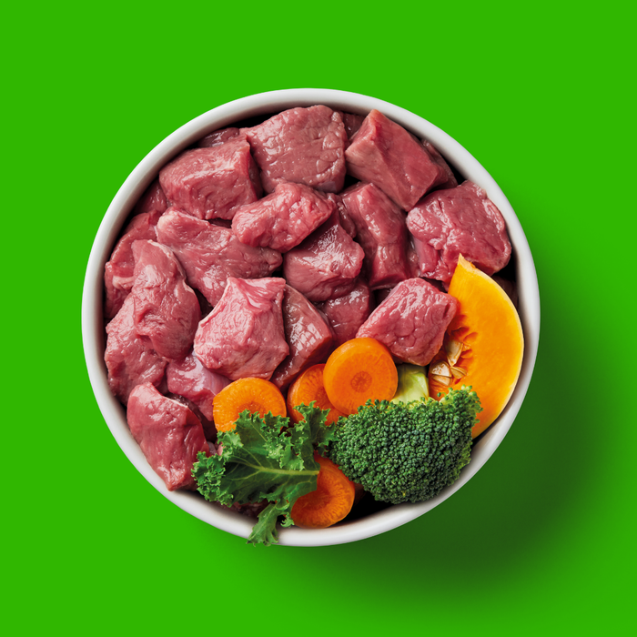 Bowl of Raw Lamb 500g cubes, broccoli, carrots, and squash on a bright green background.